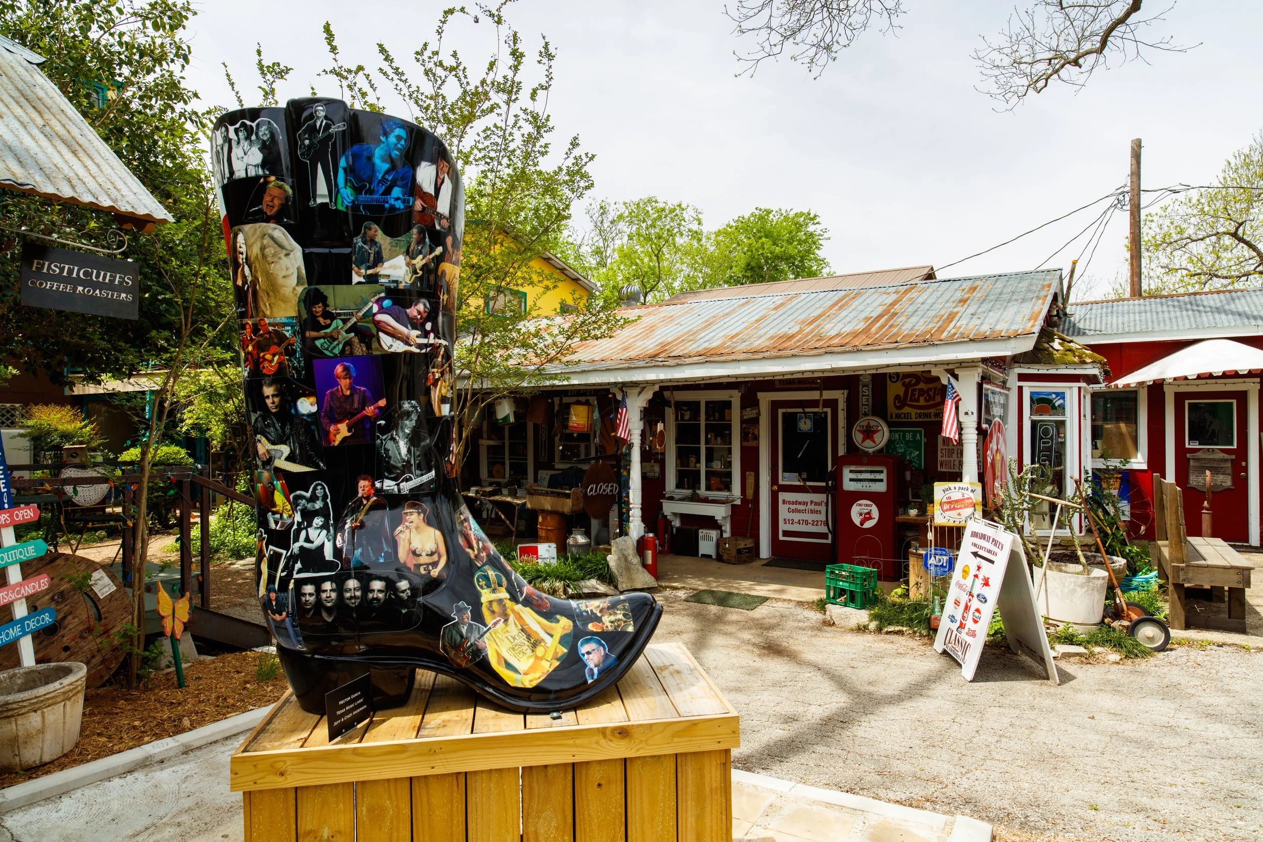A large decorative boot sculpture covered with images of musicians, placed on a wooden platform outside a rustic shop with colorful signs and an American flag.