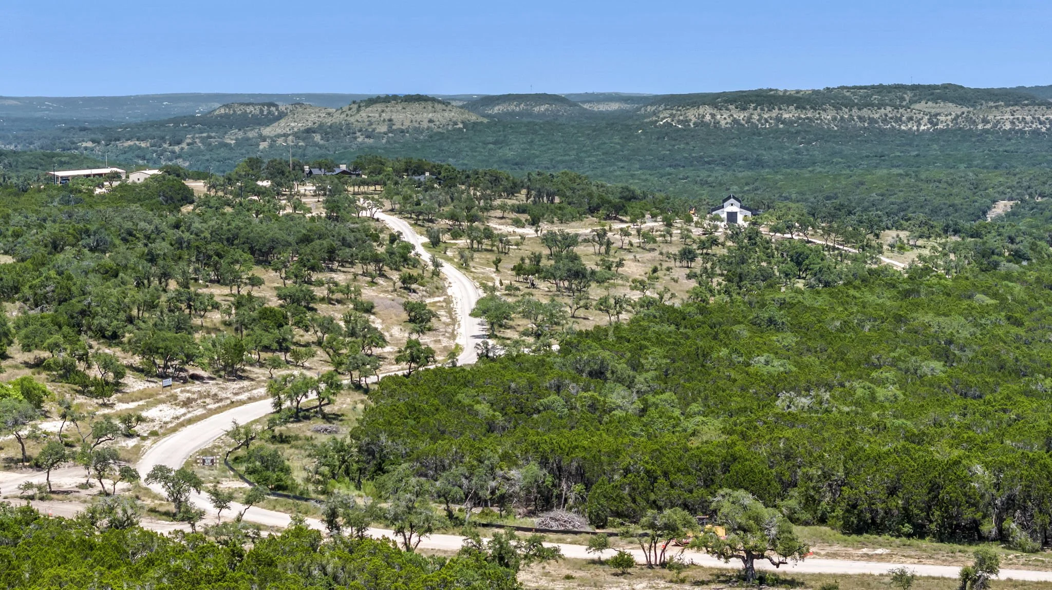 A winding dirt road running through a hilly landscape with scattered trees and a few buildings, set against distant hills under a clear blue sky.