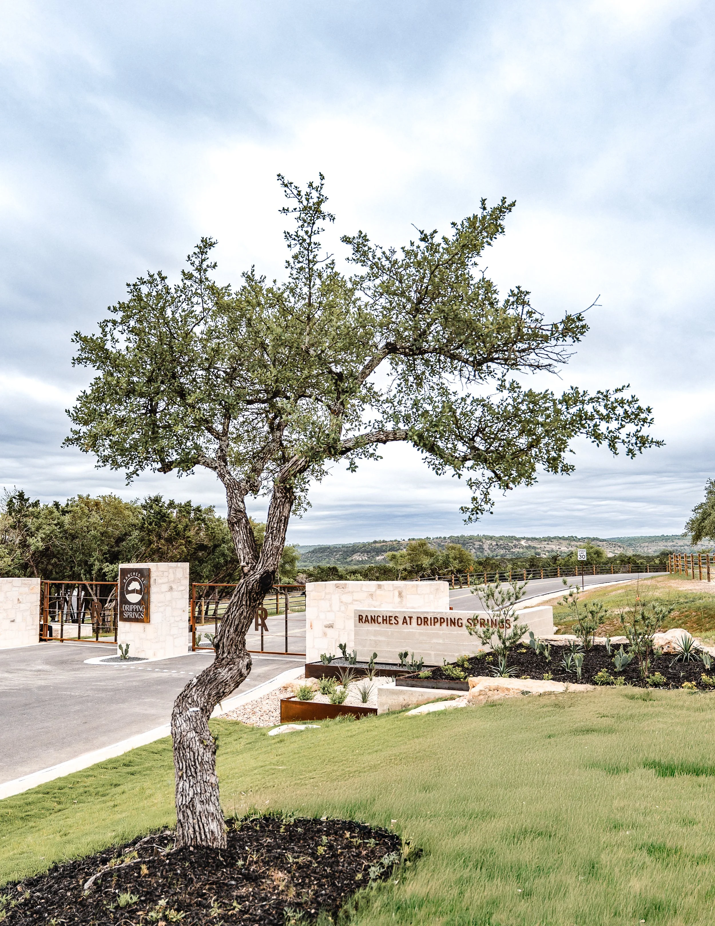 Entrance to Ranches at Dripping Springs with a curved tree, stone sign, plants, and a road in the background under cloudy sky.