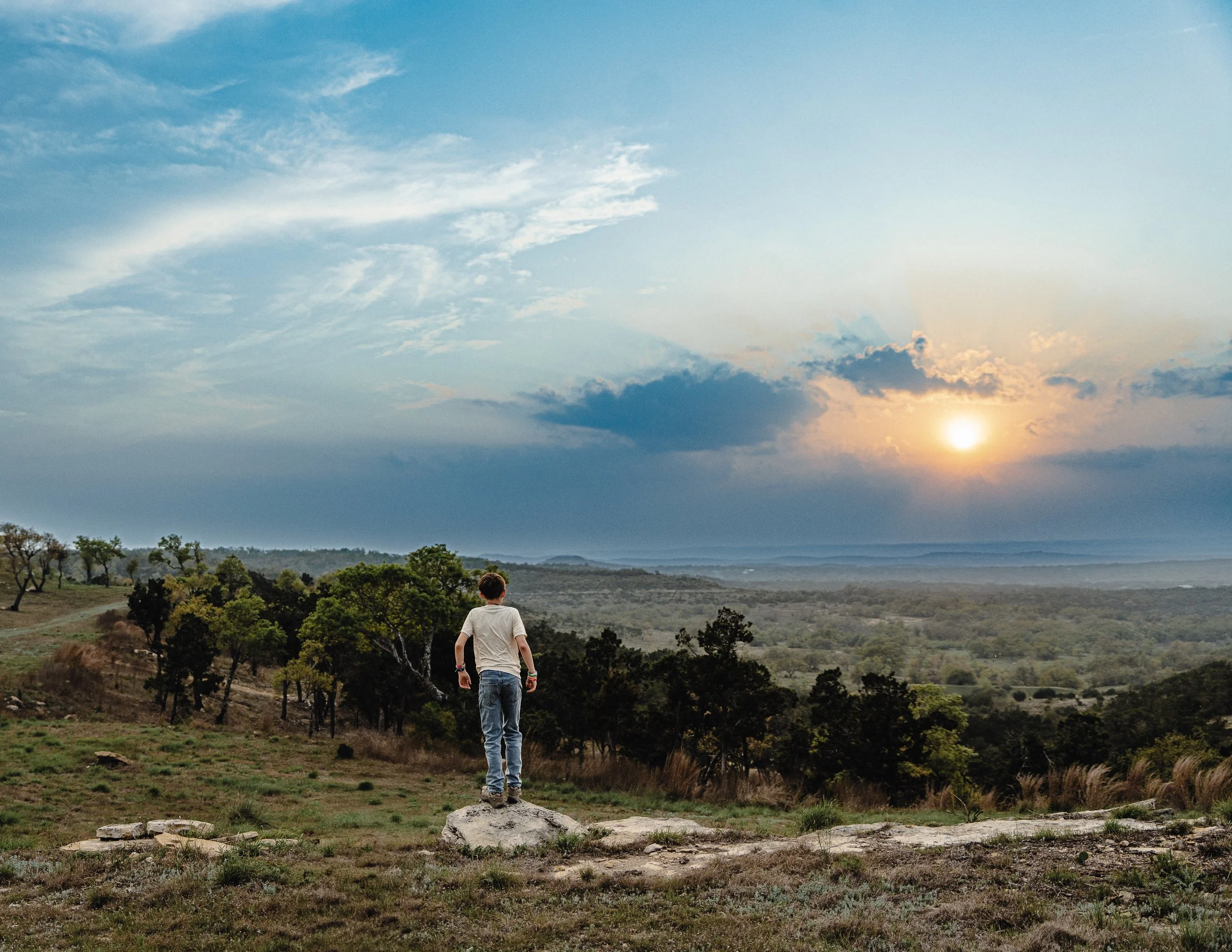 A person standing on a rock in a grassy field with trees, watching a sunset over distant hills and clouds.