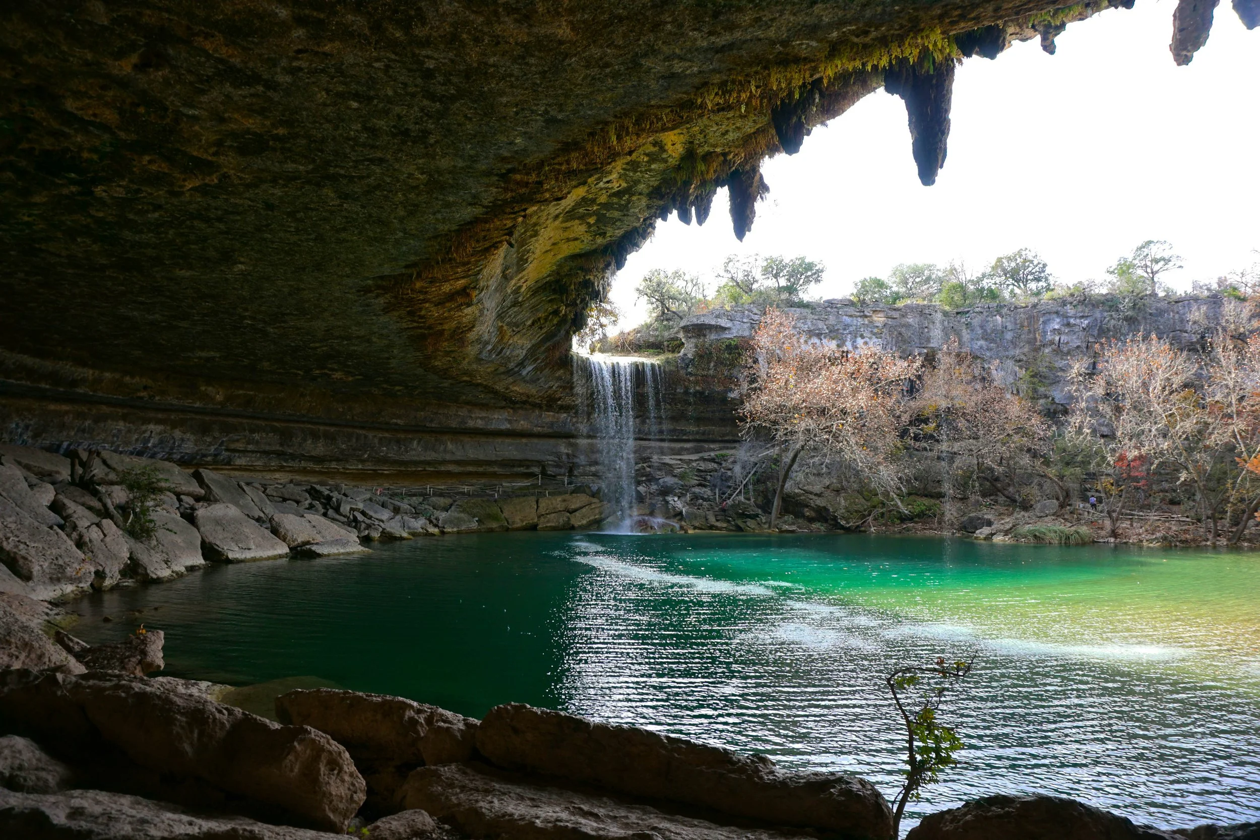 A river flowing through a rocky cave with a waterfall cascading into the water, surrounded by trees and rocky cliffs.