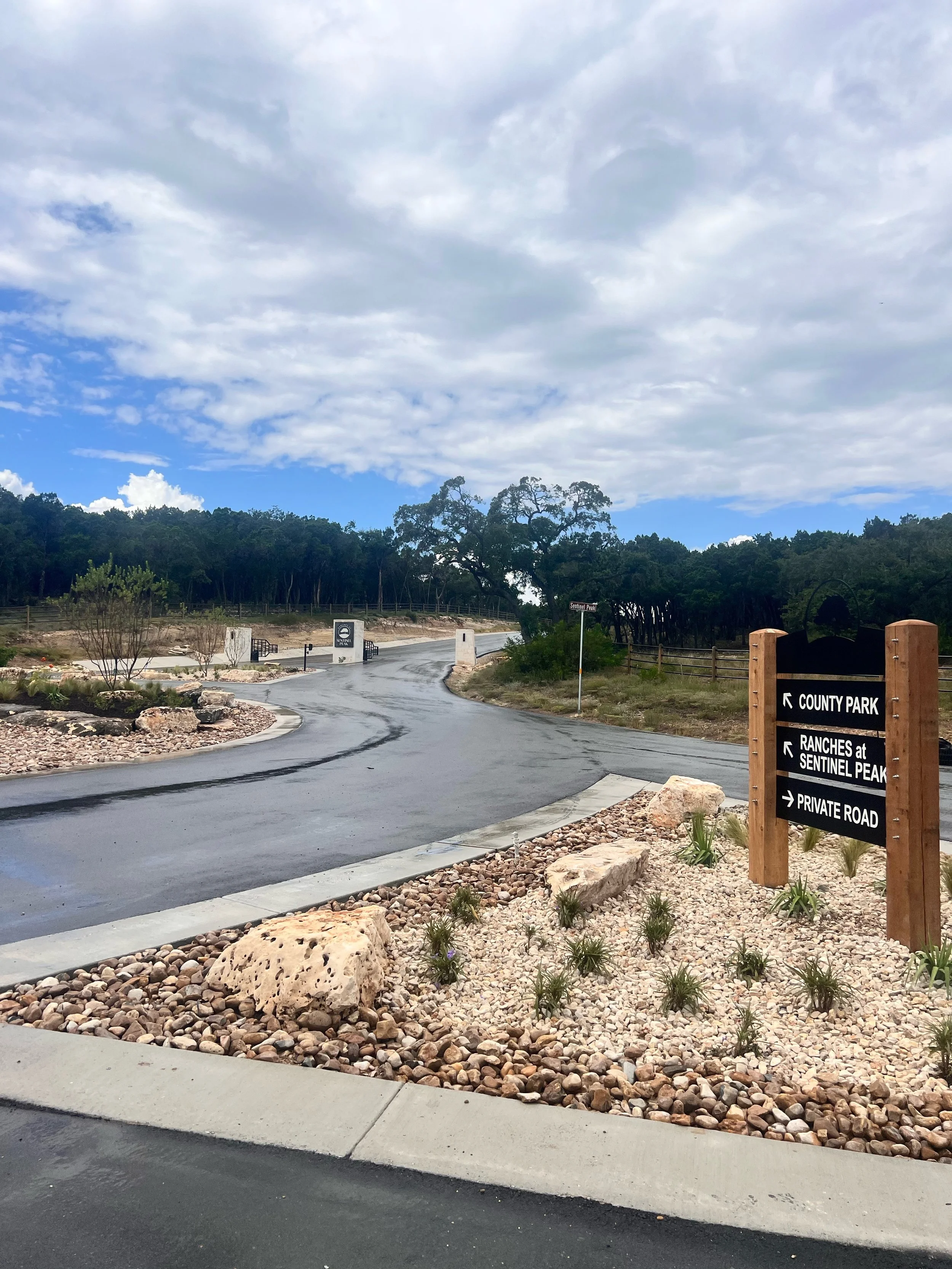 A paved road curves through a natural landscape with trees, rocks, and plants. A wooden sign indicates directions to County Park, Ranches at Sentinel Peak, and Private Road.