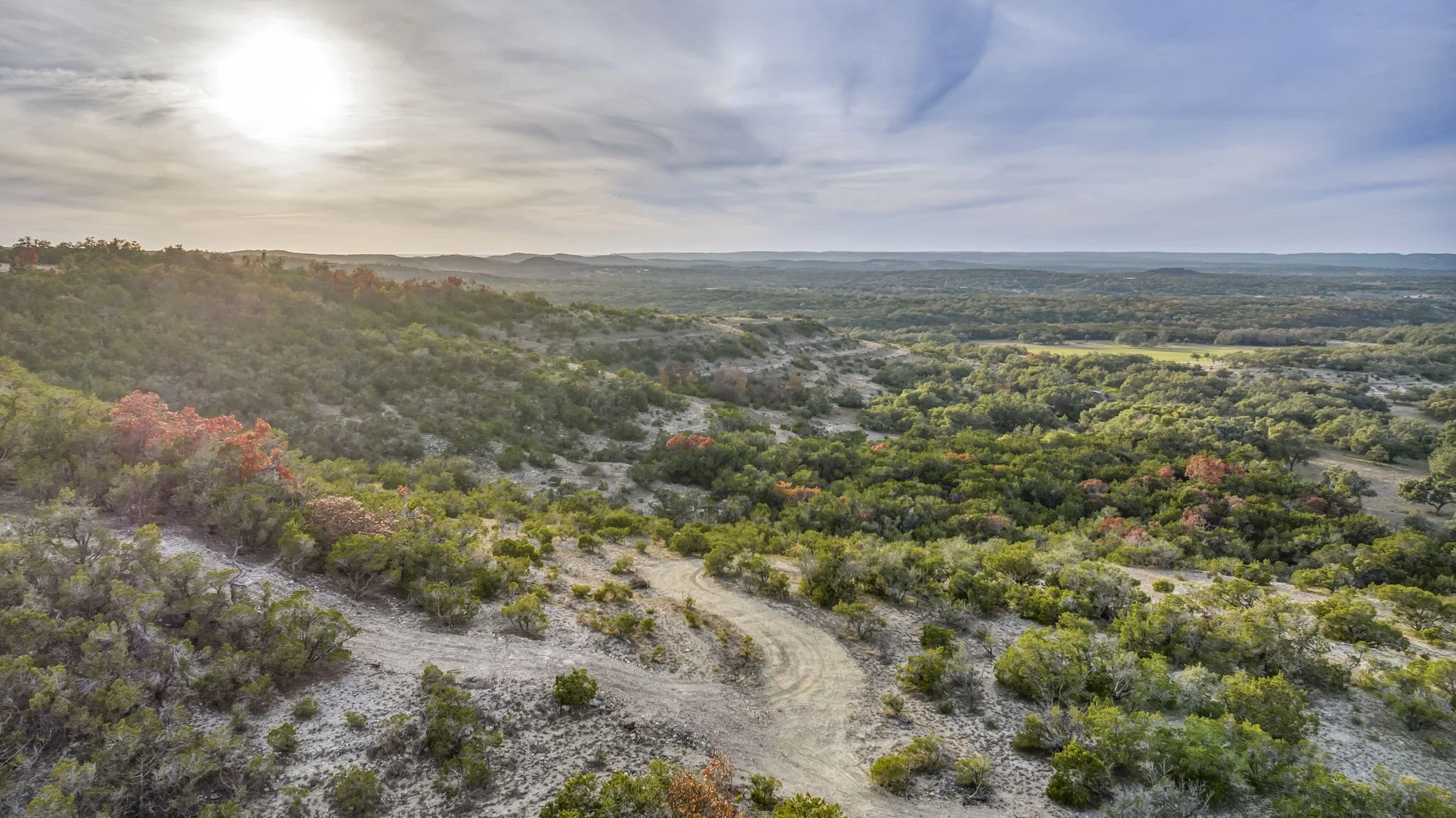 Aerial view of a vast landscape with rolling hills covered in green trees and bushes, a dirt trail winding through the terrain, under a partly cloudy sky with the sun shining through the clouds.
