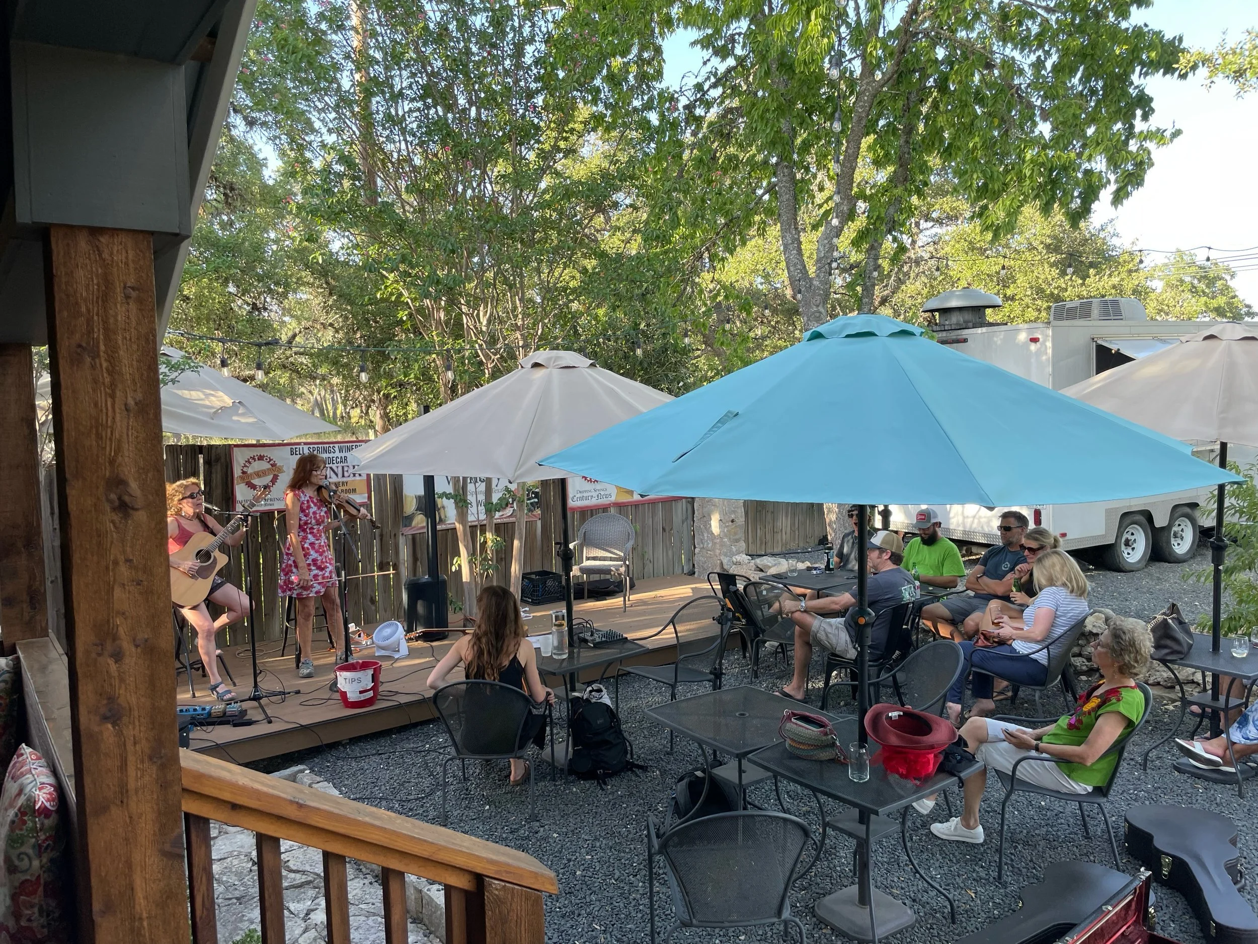 Outdoor live music performance at a casual venue, with musicians playing on a small stage and an audience sitting under large umbrellas in a shaded yard surrounded by trees.