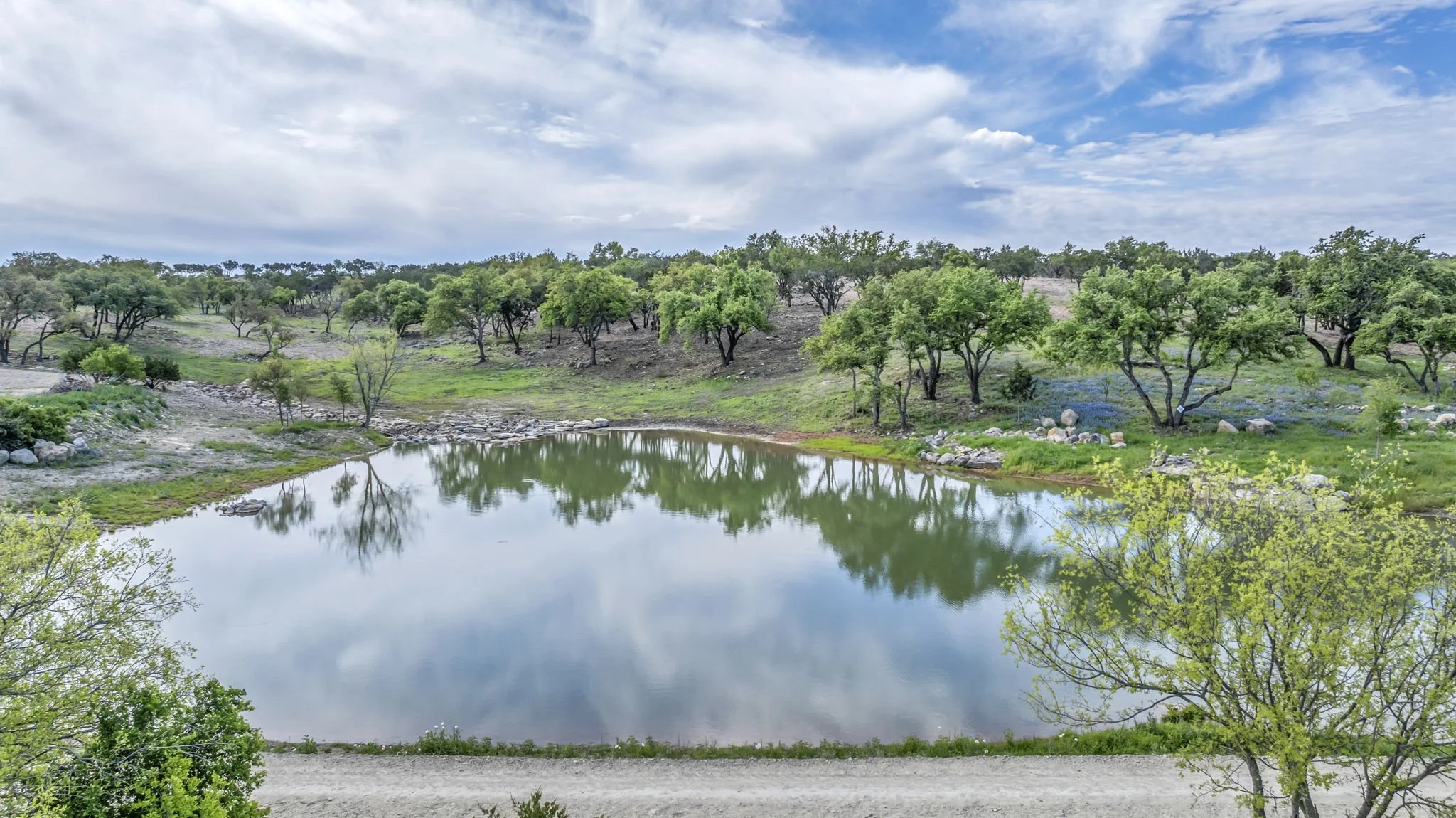 A peaceful landscape featuring a small pond reflecting the sky and surrounding green trees and hills with a partly cloudy sky.