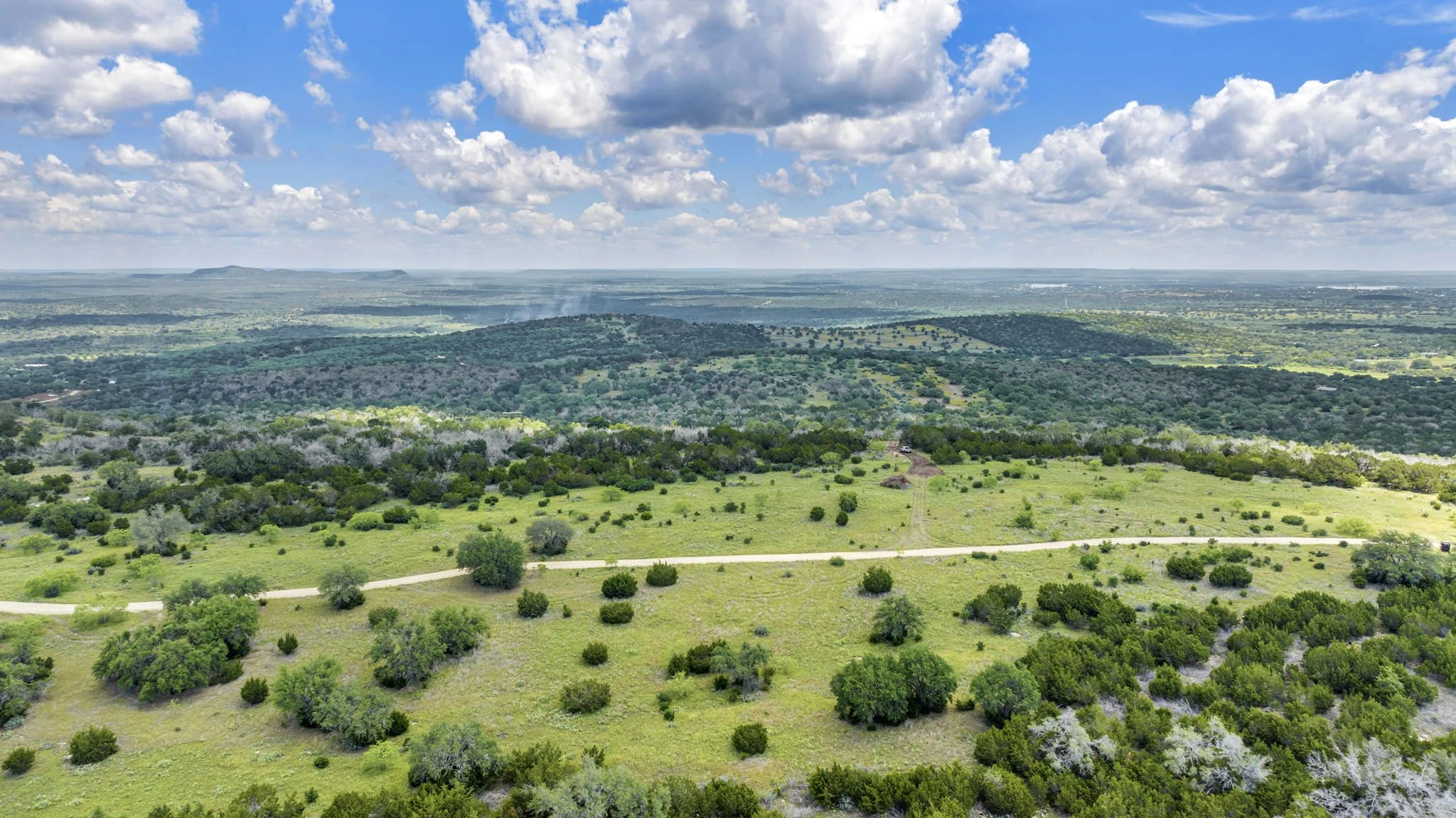 Aerial view of a vast green landscape with scattered trees, rolling hills, a dirt road, and a partly cloudy sky.