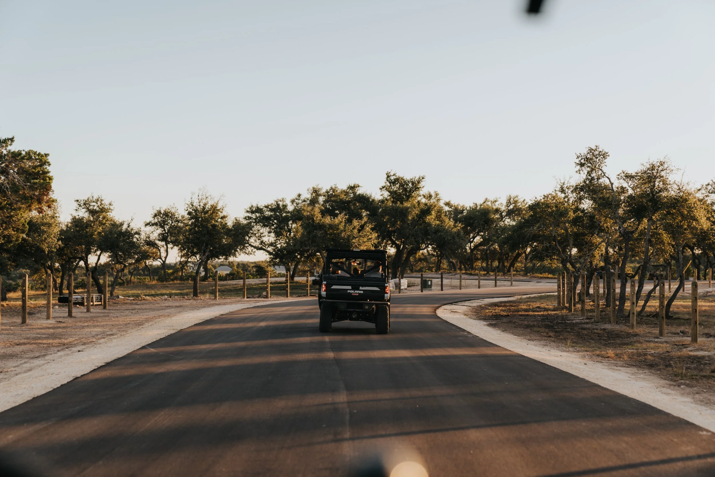 A black Polaris off-road vehicle driving on a paved winding road through a landscape with scattered trees, under a clear sky during sunset.
