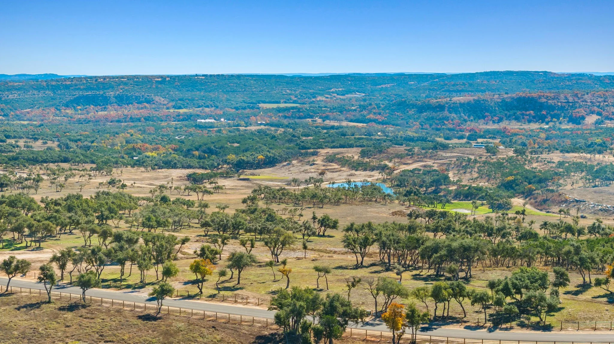 A wide landscape view of rolling hills and open fields with scattered trees under a clear blue sky, including a small pond and a paved road.