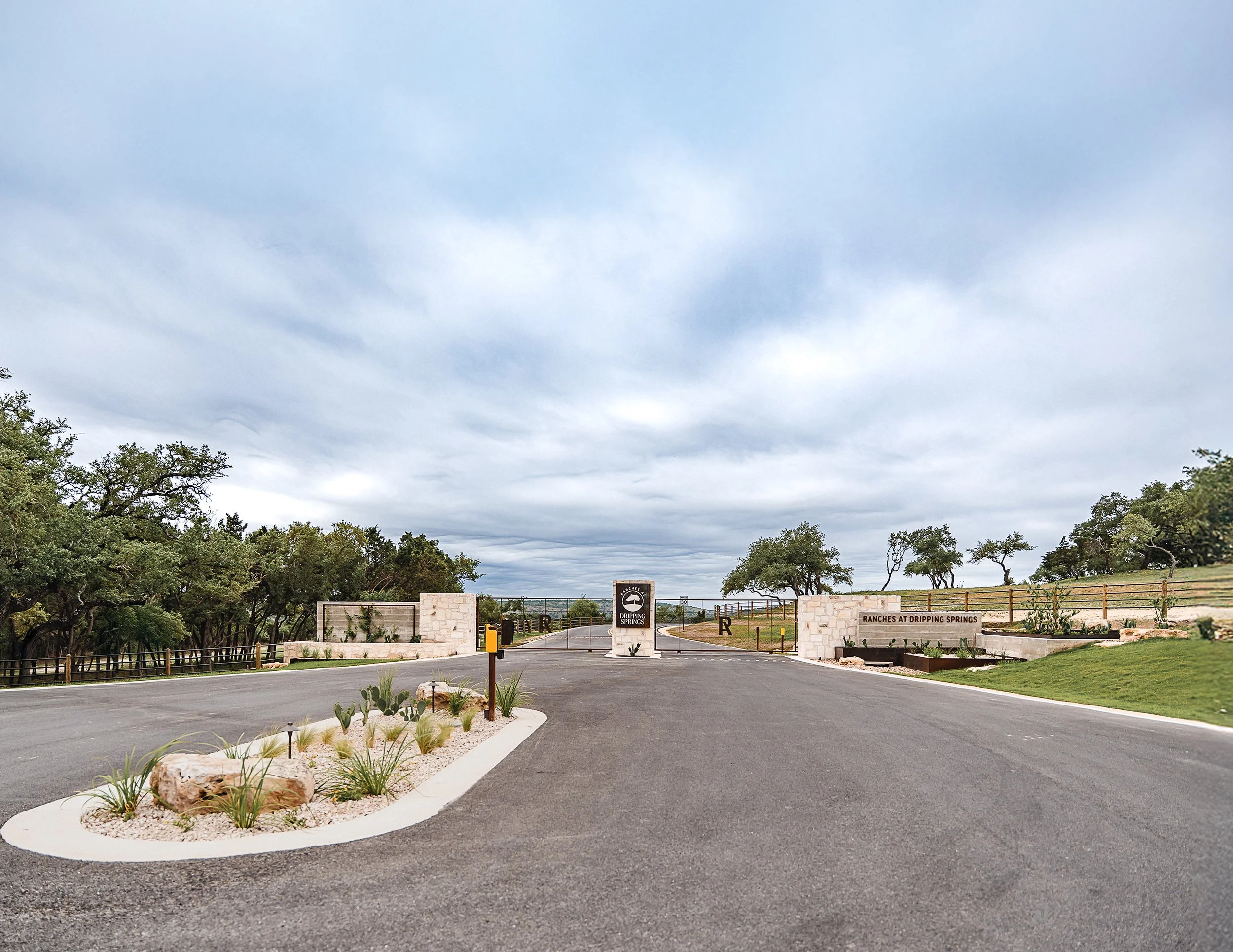 Entrance to Ranches at Dripping Springs gated community with stone markers, landscaped island, and a wide paved road, surrounded by trees and open sky.