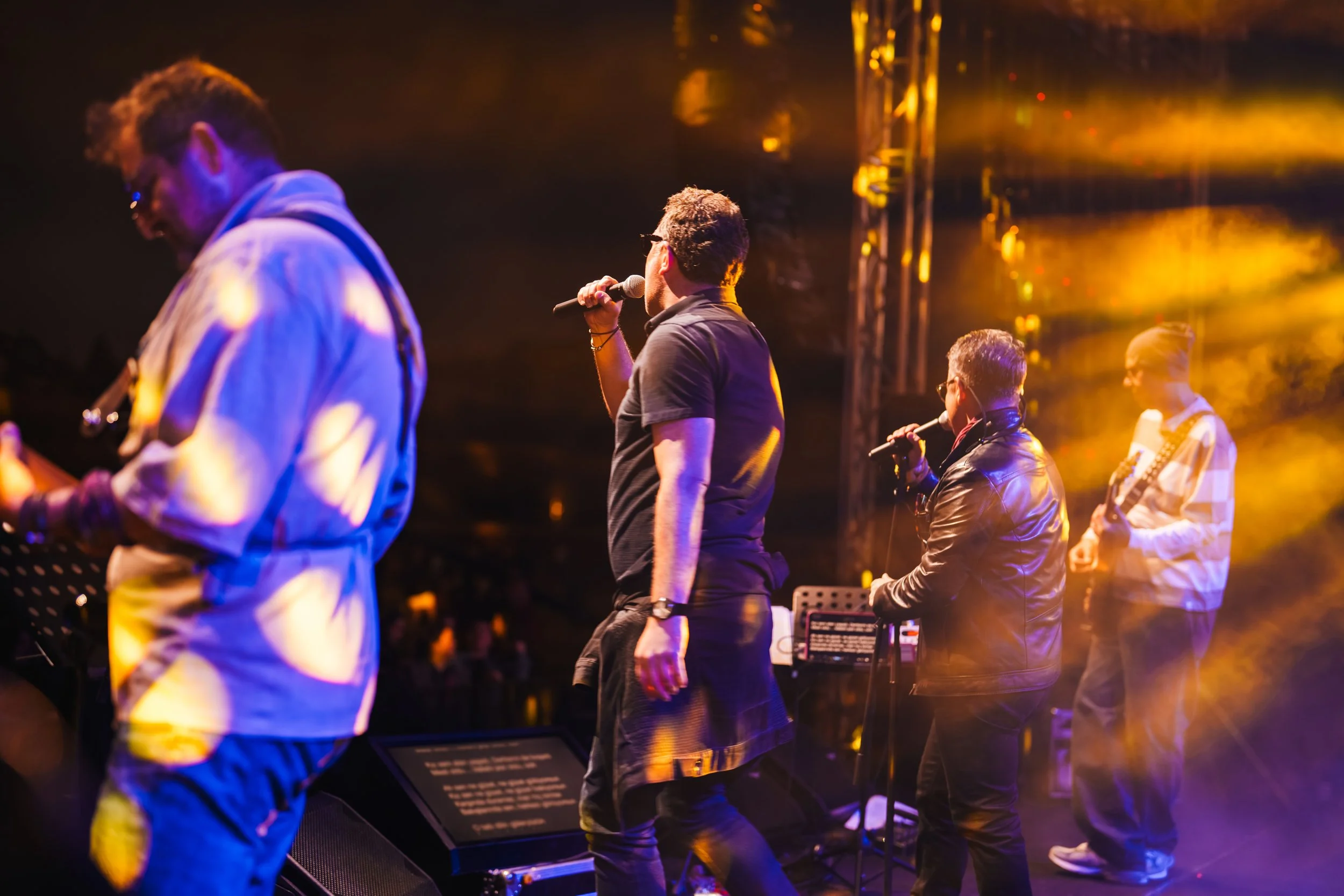 Four musicians performing on stage at night, with colorful stage lighting and equipment, including microphones and a music stand.