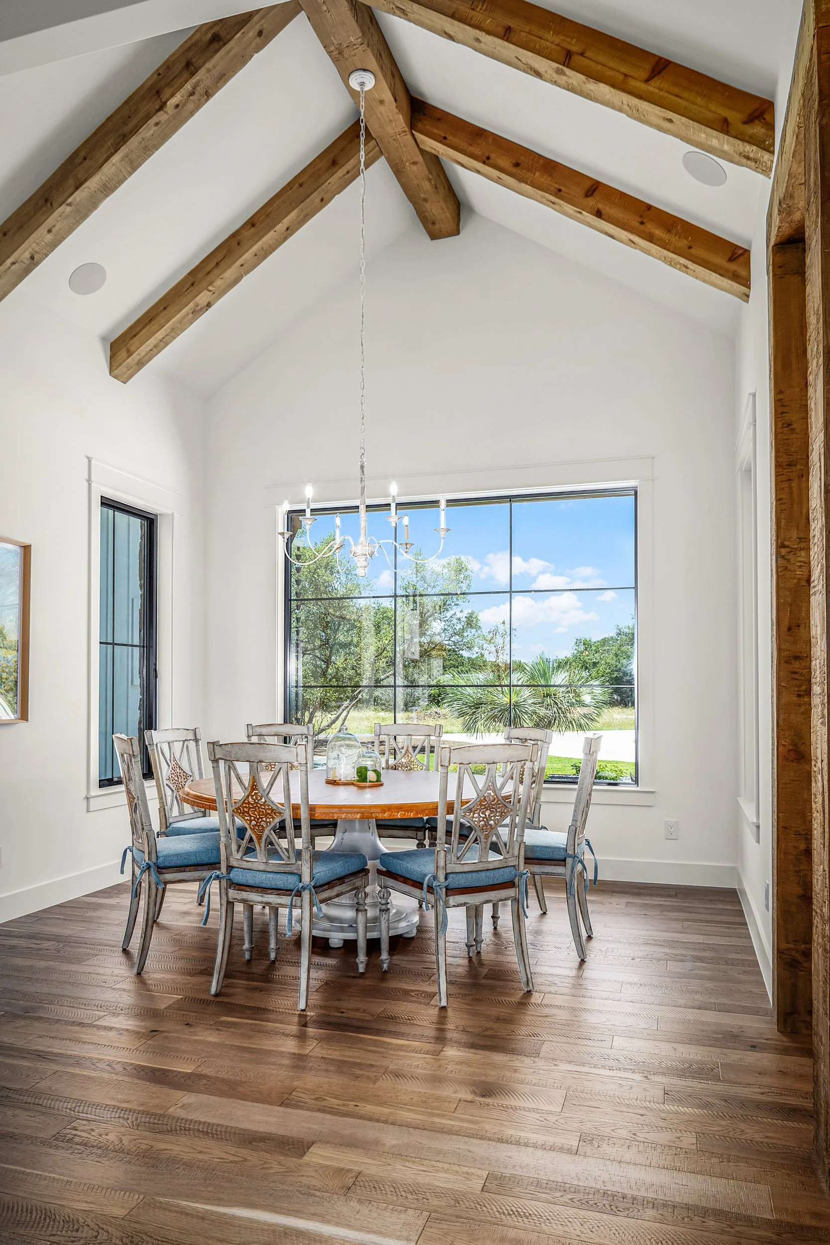 A dining room with wooden flooring, white walls, a large window showing greenery outside, a round table with eight chairs, a chandelier hanging above, and exposed wooden beams on the ceiling.