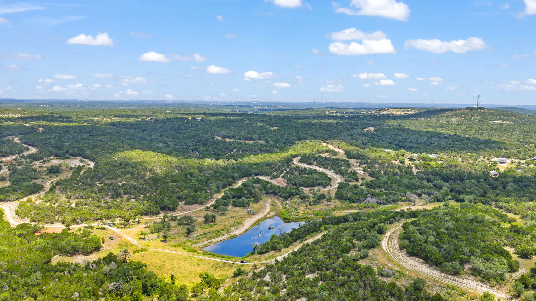 Aerial view of a green, semi-arid landscape with scattered trees and patches of bare soil, under a blue sky with some clouds.