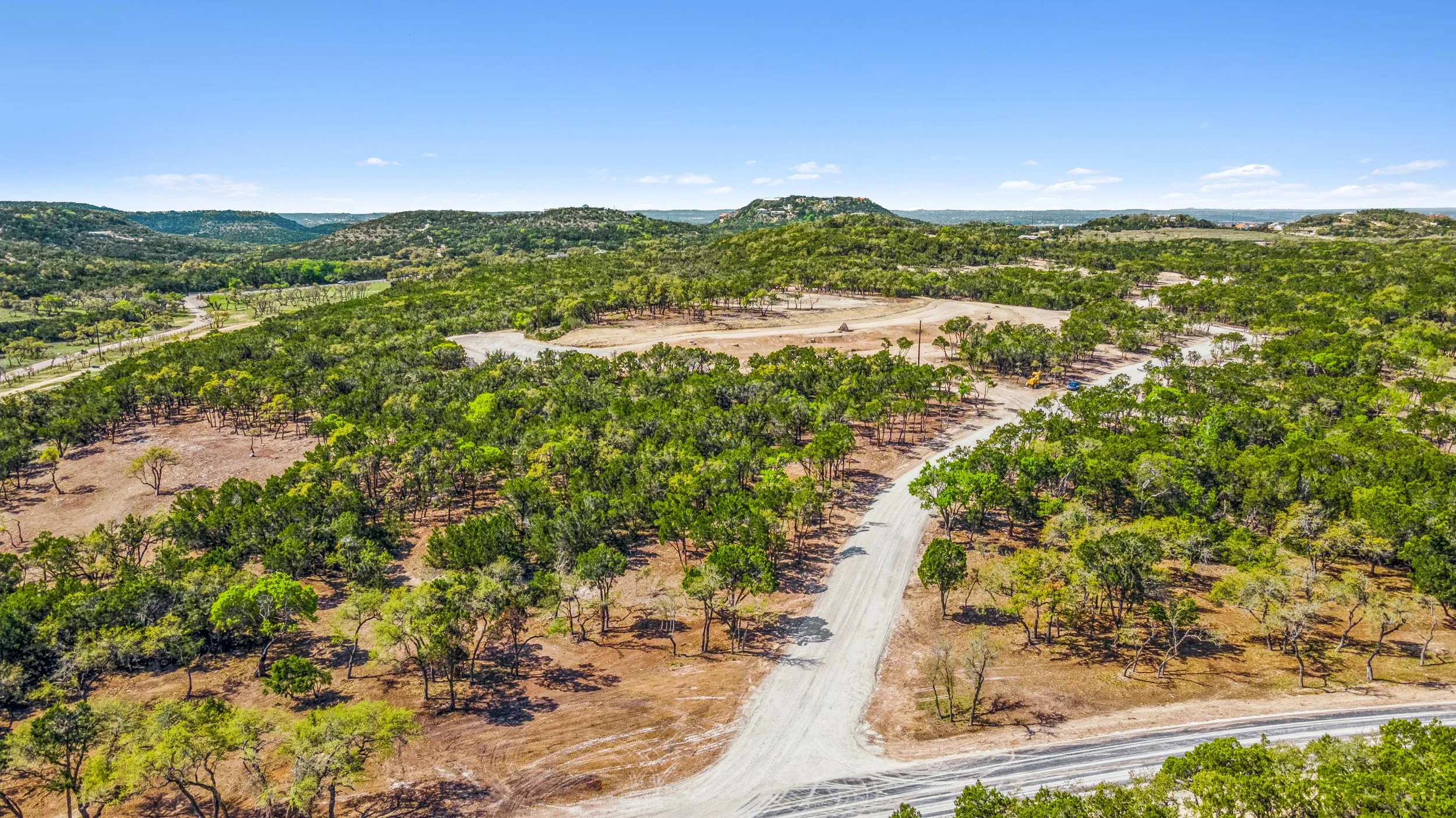 Aerial view of a green and brown landscape with scattered trees and dirt roads under a clear blue sky.